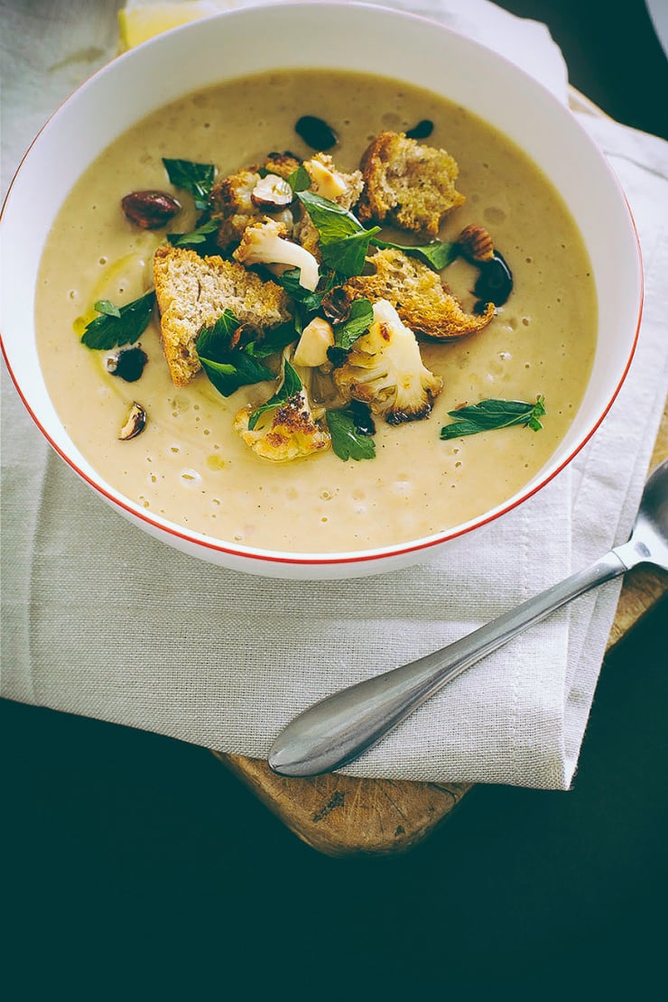 An overhead shot of a bowl of vegan roasted cauliflower soup, garnished with croutons and chopped parsley. The soup is beige. The bowl is on a beige napkin, which is on top of a wooden cutting board.