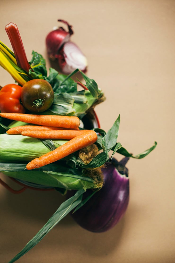An overhead portrait shot of prepped summer vegetables including sliced eggplant, tomatoes, corn, and chard arranged on a surface ready for cooking.