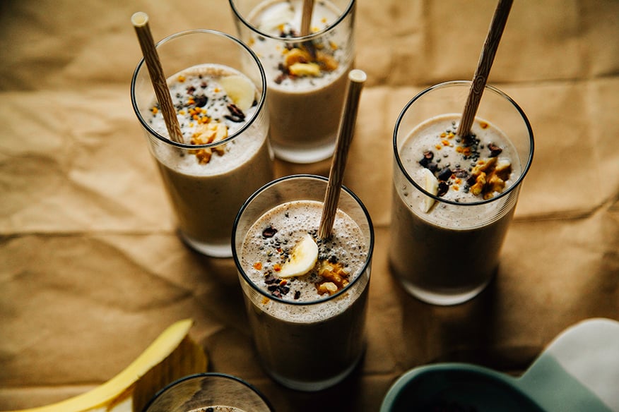 An overhead shot of several banana bread shakes in slim glasses on a brown paper background. Shakes are garnished with sliced banana, walnuts, and cacao nibs and finished with beige paper straws.