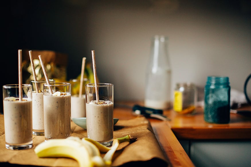 An head-on shot of several banana bread shakes in slim, clear glasses on a brown paper background. Shakes are garnished with sliced banana, walnuts, and cacao nibs and finished with beige paper straws.