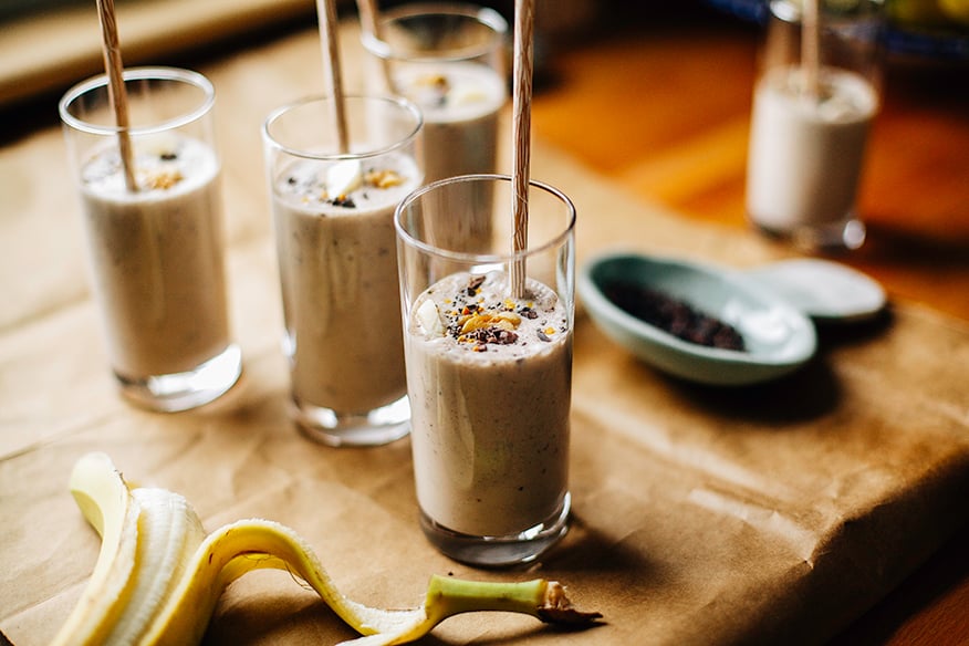 An head-on shot of several banana bread shakes in slim, clear glasses on a brown paper background. Shakes are garnished with sliced banana, walnuts, and cacao nibs and finished with beige paper straws.