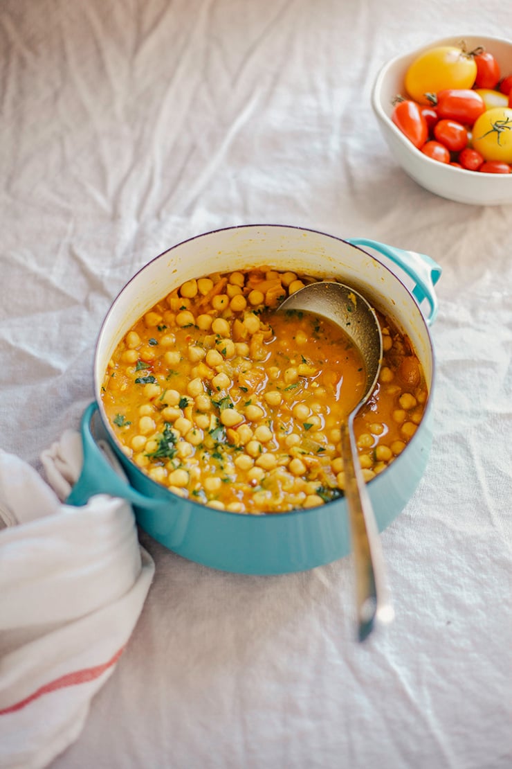 A 3/4 angle shot shows a pot of golden chana masala in a small turquoise pot. A small bowl of tomatoes is seen nearby and a ladle is sticking out of the pot.