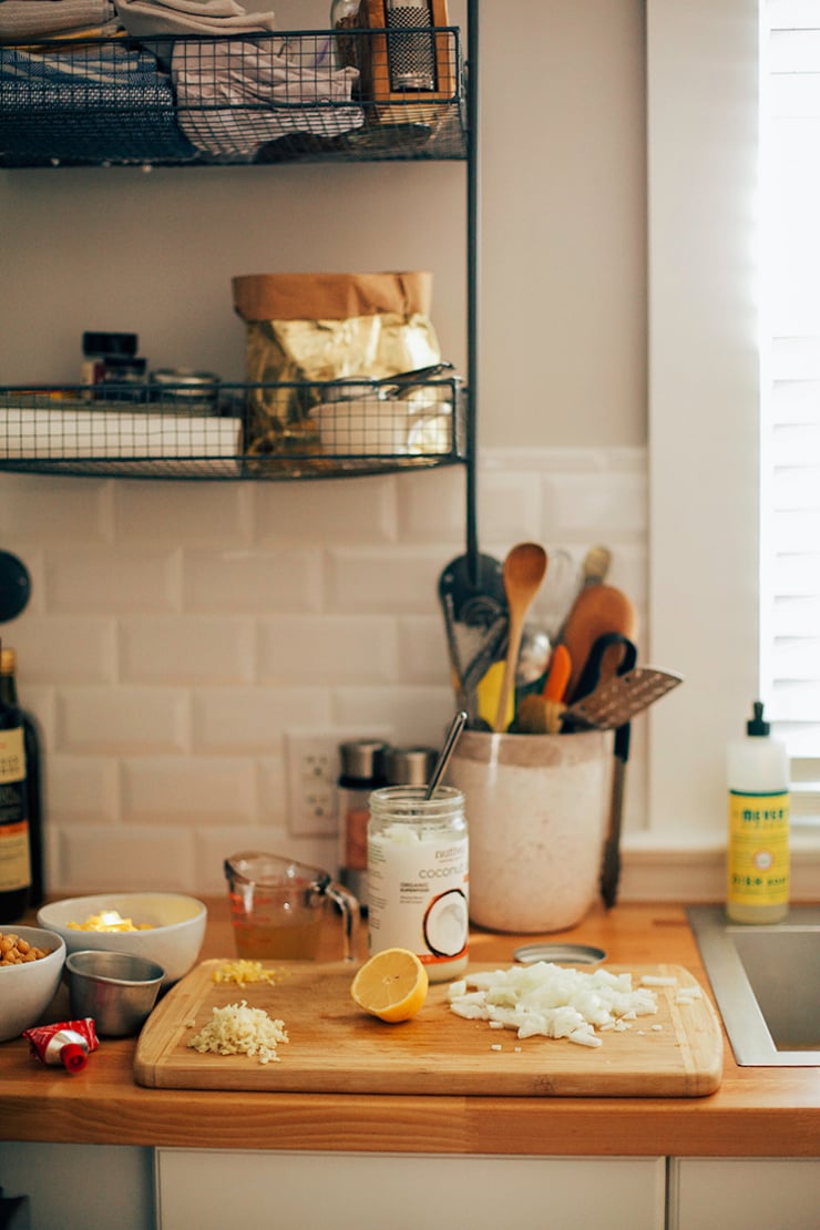 A head-on kitchen counter scene shoes a cutting board with chopped onions, minced garlic and minced ginger on top. A jar of coconut oil and a vase filled with kitchen cooking utensils  is also visible.