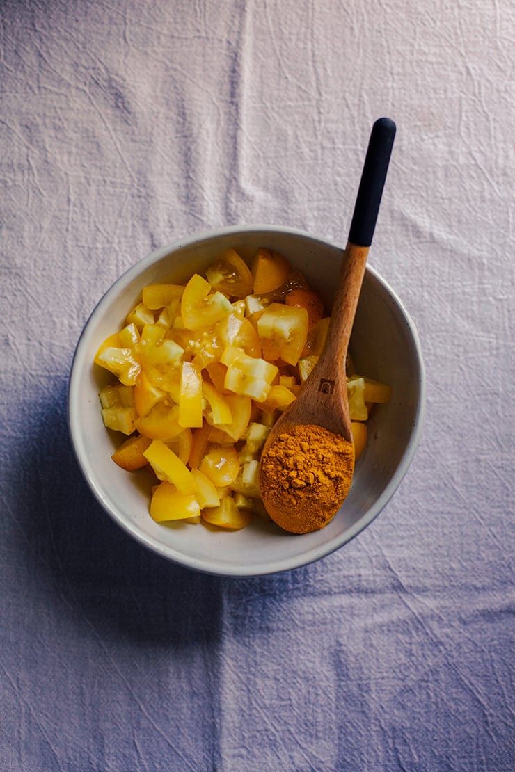 An overhead shot shows a bowl of chopped yellow tomatoes and a spoonful of turmeric resting in the bowl.