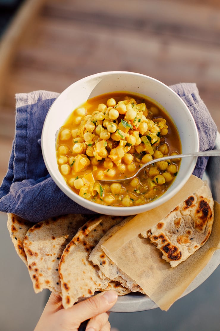 An overhead shot shows a hand holding a plate that contains a bowl of golden chana masala, a linen napkin, and torn pieces of whole grain flatbread.