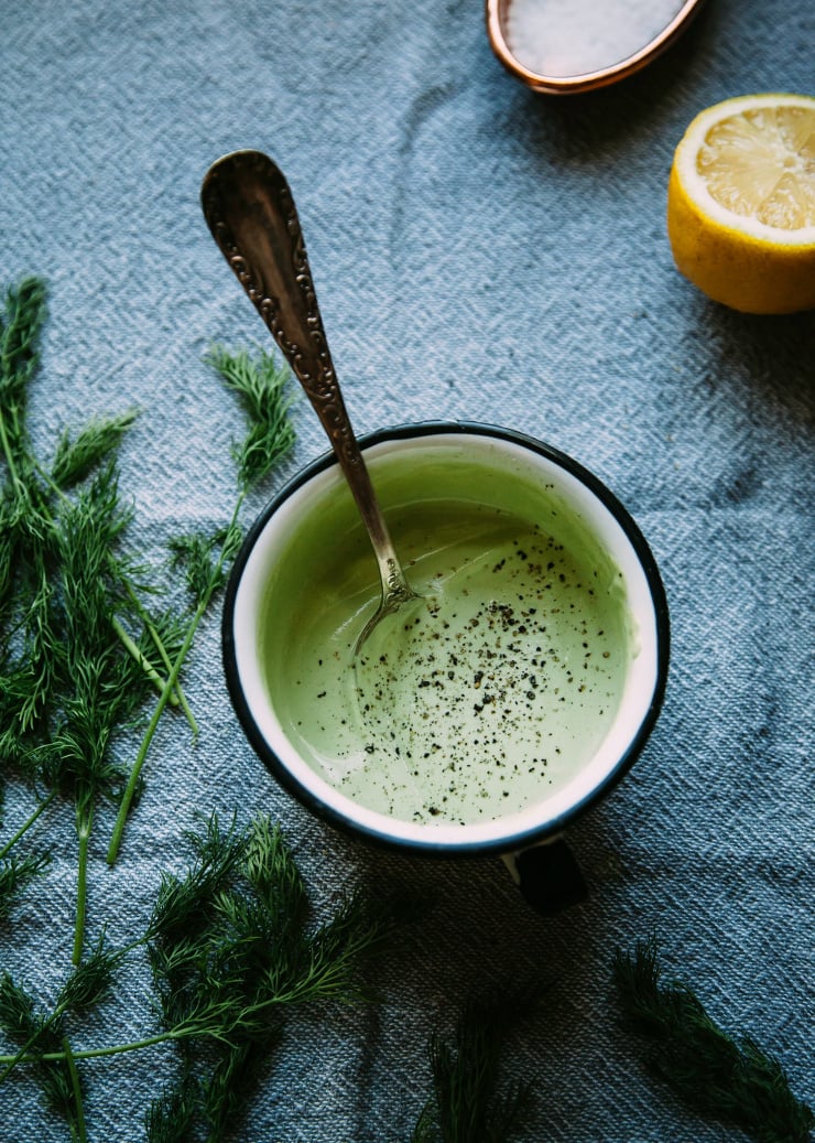 An overhead shot of a cup of light green, creamy sauce in a white enamelware mug. A halved lemon is seen to the side.