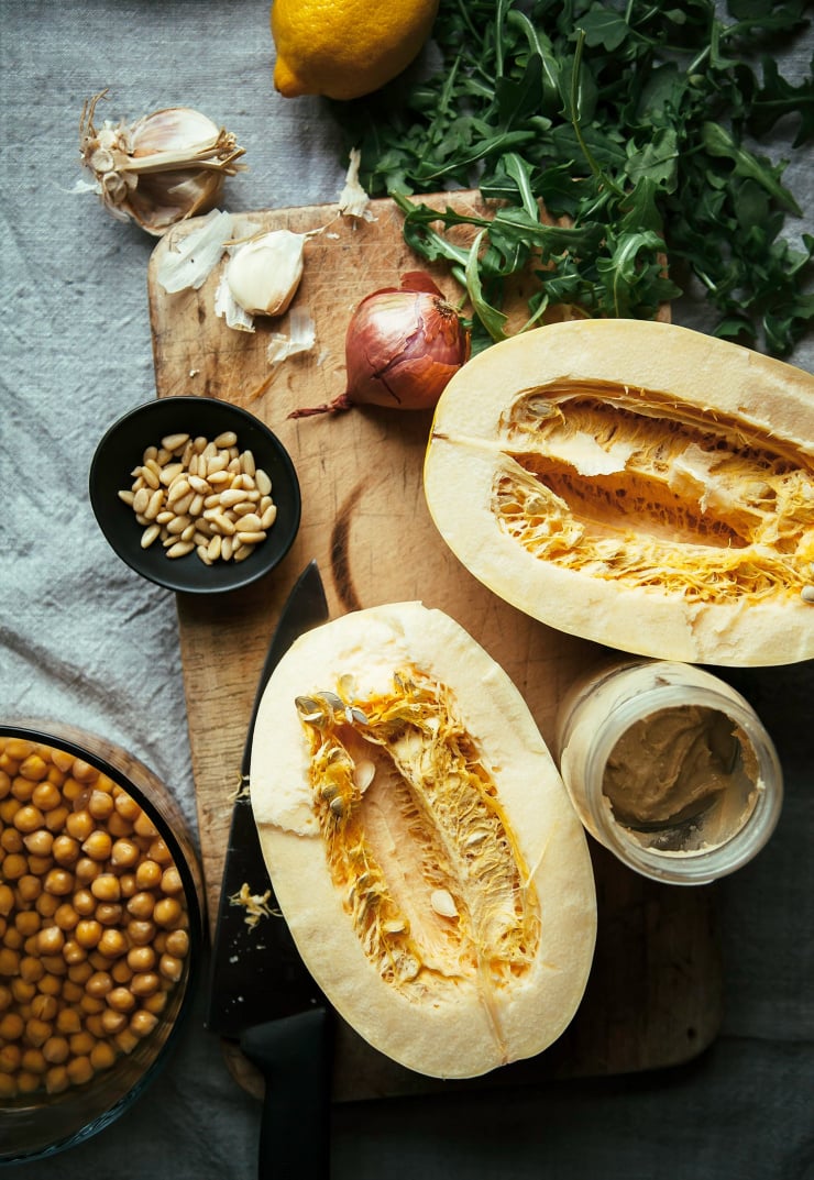 An overhead shot of ingredients for stuffed spaghetti squash on top of a worn wooden cutting board that&rsquo;s set on top of a grey linen tablecloth.