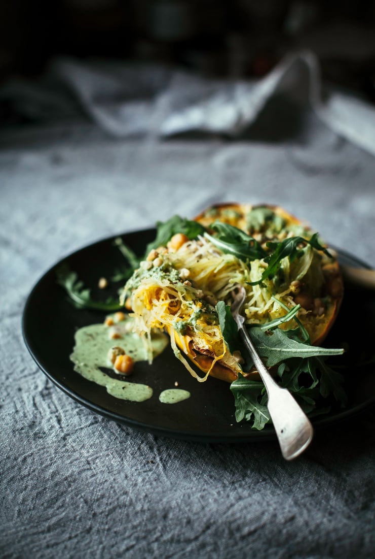 A side angle of a stuffed spaghetti squash with arugula cream and chickpeas. A fork is sticking out. The squash is on a black plate on top of a grey linen tablecloth.