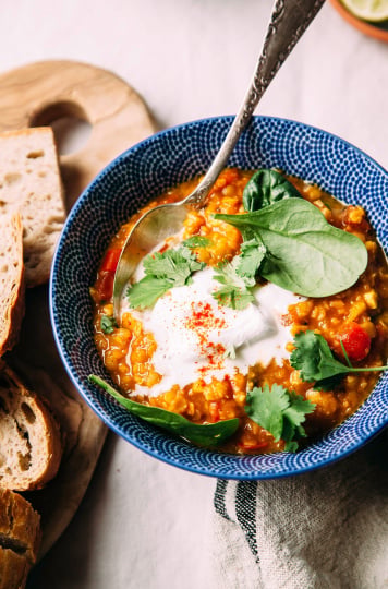 Golden lentil soup with coconut spinach and lime, photographed from overhead in a deep blue bowl, garnished with a dollop of coconut yogurt. There is a wood board with sliced bread nearby. All is photographed on a beige linen tablecloth.