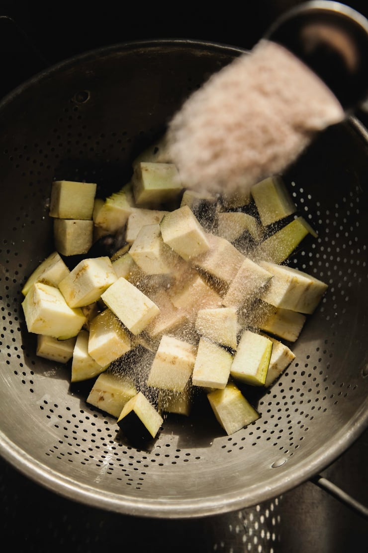 An overhead shot shows cubed eggplant in a colander with salt being poured on top.
