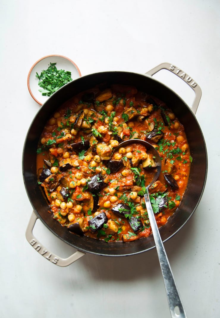 An overhead shot shows a pot filled with braised harissa eggplant with chickpeas. The dish is garnished with chopped parsley. A ladle sticks out of the pot. A small bowl of chopped parsley is seen to the side.
