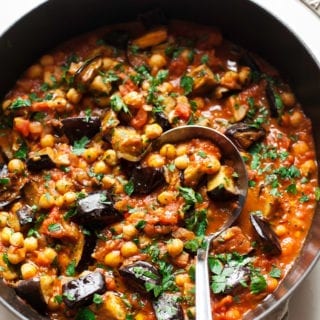 An overhead shot of a stew in a braiser-style pot, containing: seared chunks of eggplant, chickpeas, crushed tomatoes, spices, and chopped parsley.