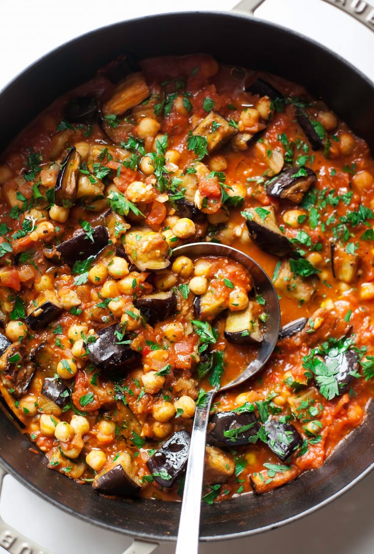 An up close, overhead shot shows a pot filled with braised harissa eggplant with chickpeas. The dish is garnished with chopped parsley. A ladle sticks out of the pot.