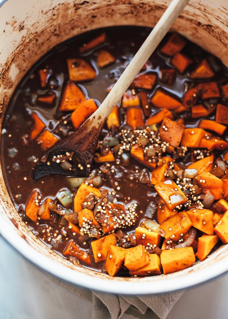 An overhead shot shows a pot filled with cubed butternut squash, quinoa, and a dark, spice and cocoa-imbued broth.