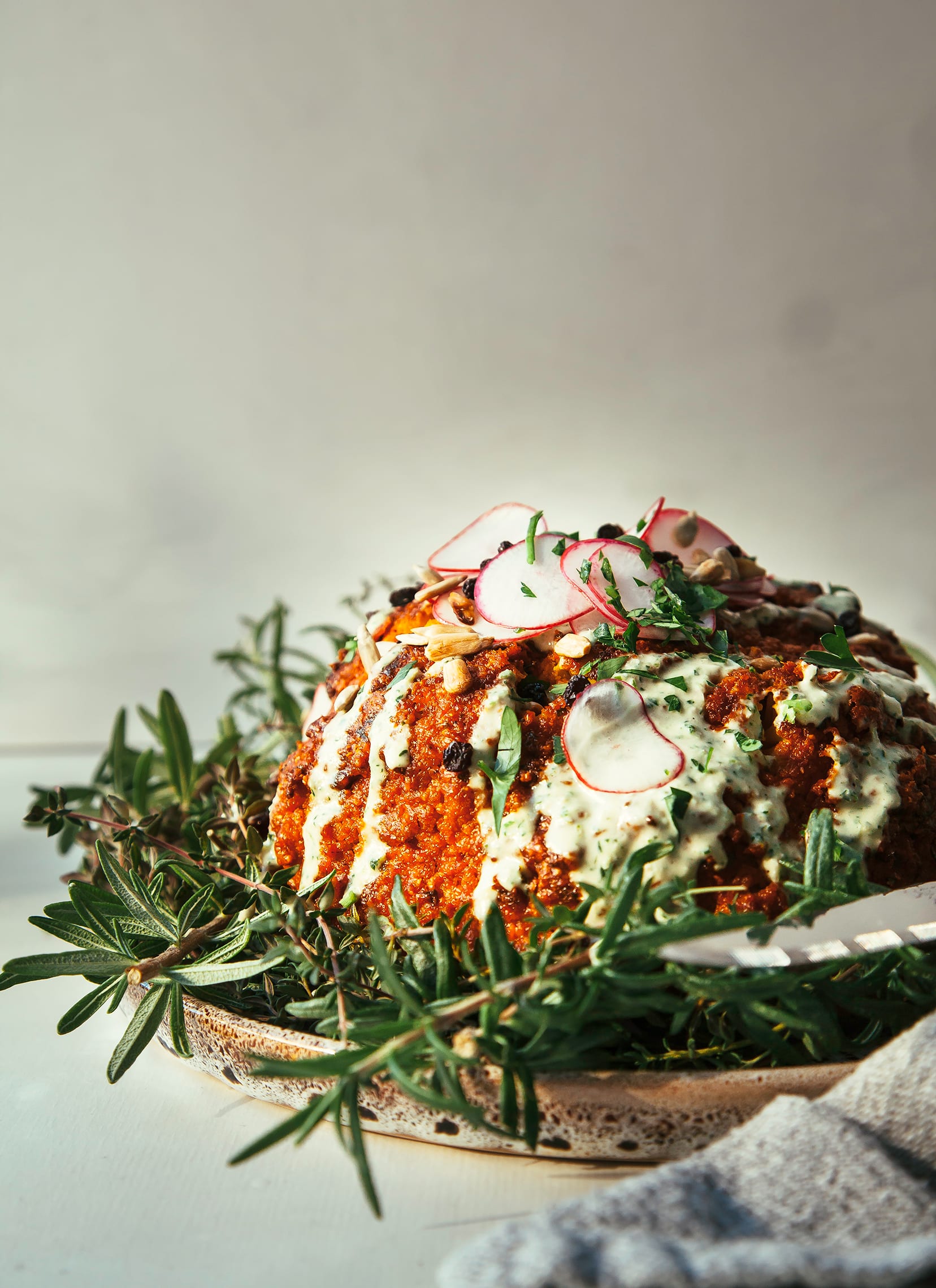 Image shows a whole roasted cauliflower coated in deep red romesco sauce. The cauliflower is garnished with a creamy light green sauce and sliced radishes.