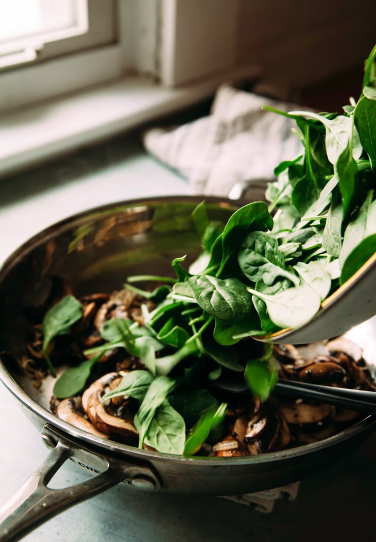 Image shows baby spinach being added to a saut&eacute; pan with mushrooms.
