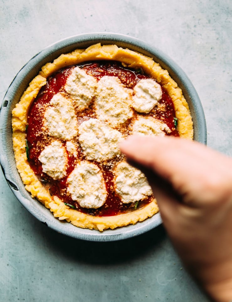 Image shows a hand sprinkling a vegan &ldquo;Parmesan&rdquo; over a filled polenta pie.