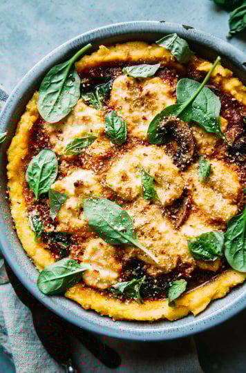 An overhead shot of a baked savoury polenta pie filled with cooked mushrooms, wilted spinach, dollops of almond-based "ricotta" and marinara sauce.