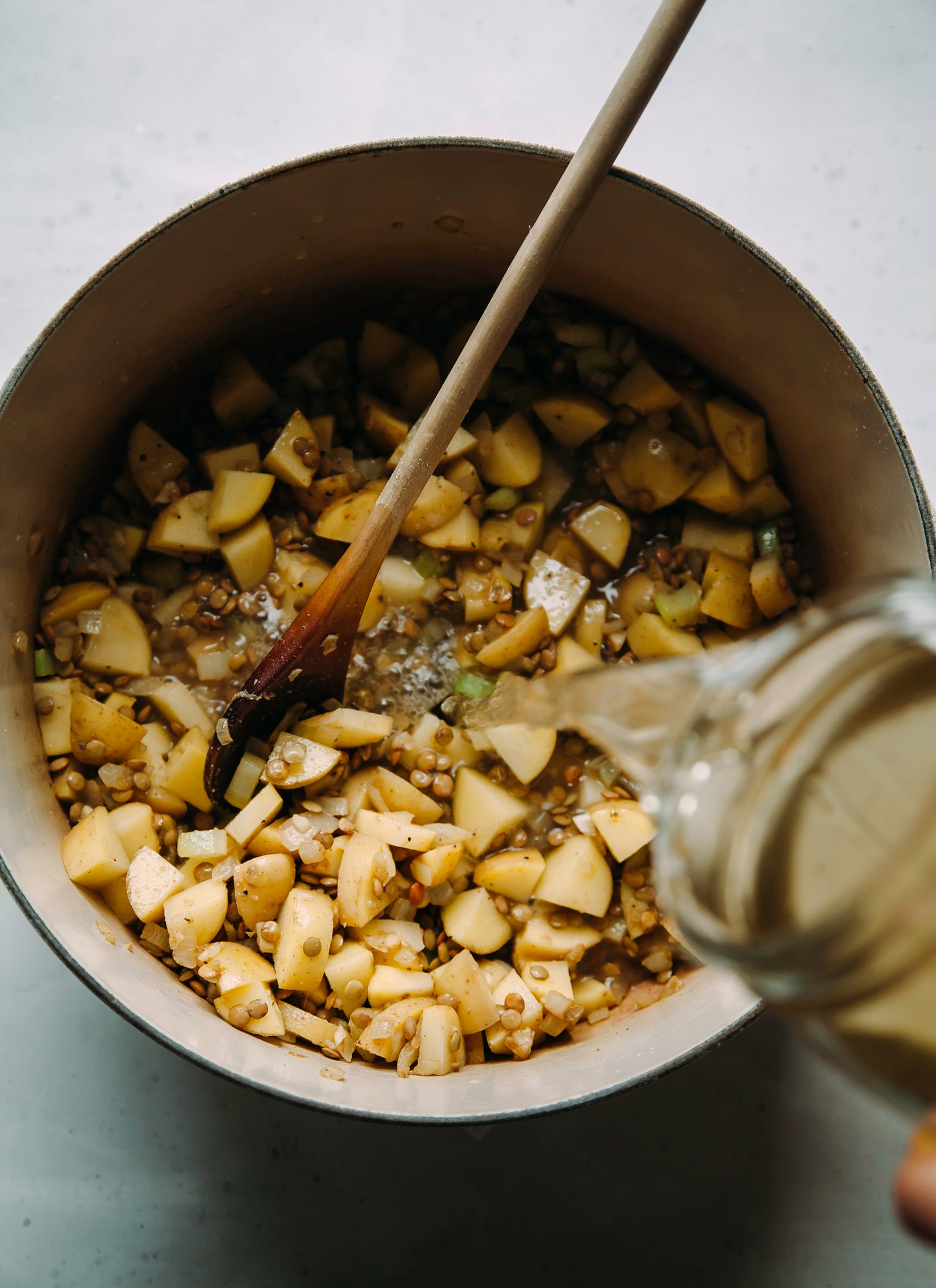 A hand is pouring vegetable stock into a soup pot filled with potatoes and lentils.