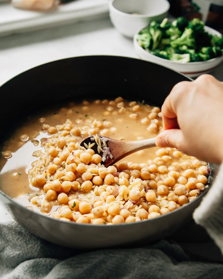 Image shows a hand storring chickpeas in a braiser-style pot. The image is taken at a 3/4 angle and the chickpeas are slightly submerged in broth.