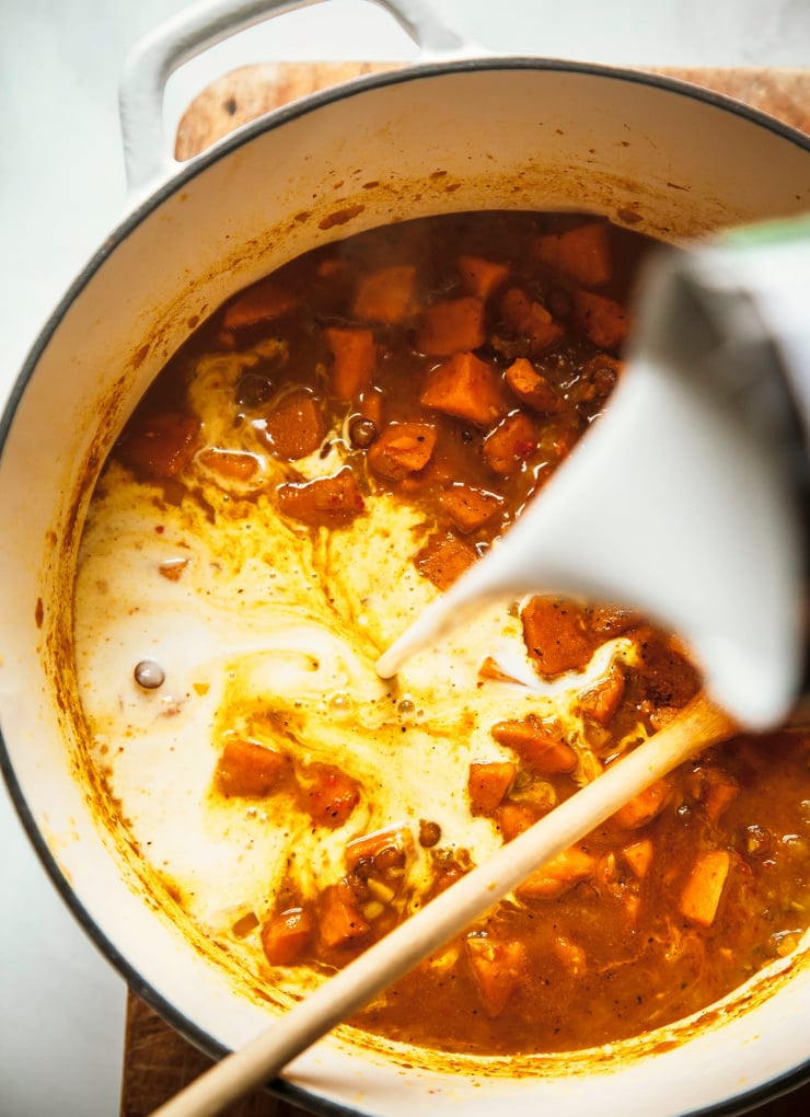 An up close, overhead shot of coconut milk being poured into a pot of soup with lentils and spices.