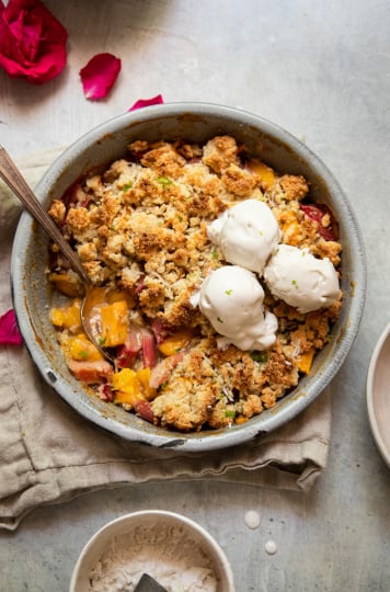 An overhead shot of a rhubarb mango crumble in a grey enamelware, round dish. It has a crisp, beige cookie-like topping. The crumble has two melt-y scoops of ice cream on top.