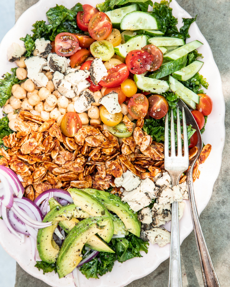 An overhead shot of a composed salad in bright, outdoor light. Salad is photographed on a link plate and includes: halved cherry tomatoes, kale, sliced cucumber, sliced avocado, chickpeas, toasted almond "bacon", sliced red onion, and bits of crumbled vegan cheese.