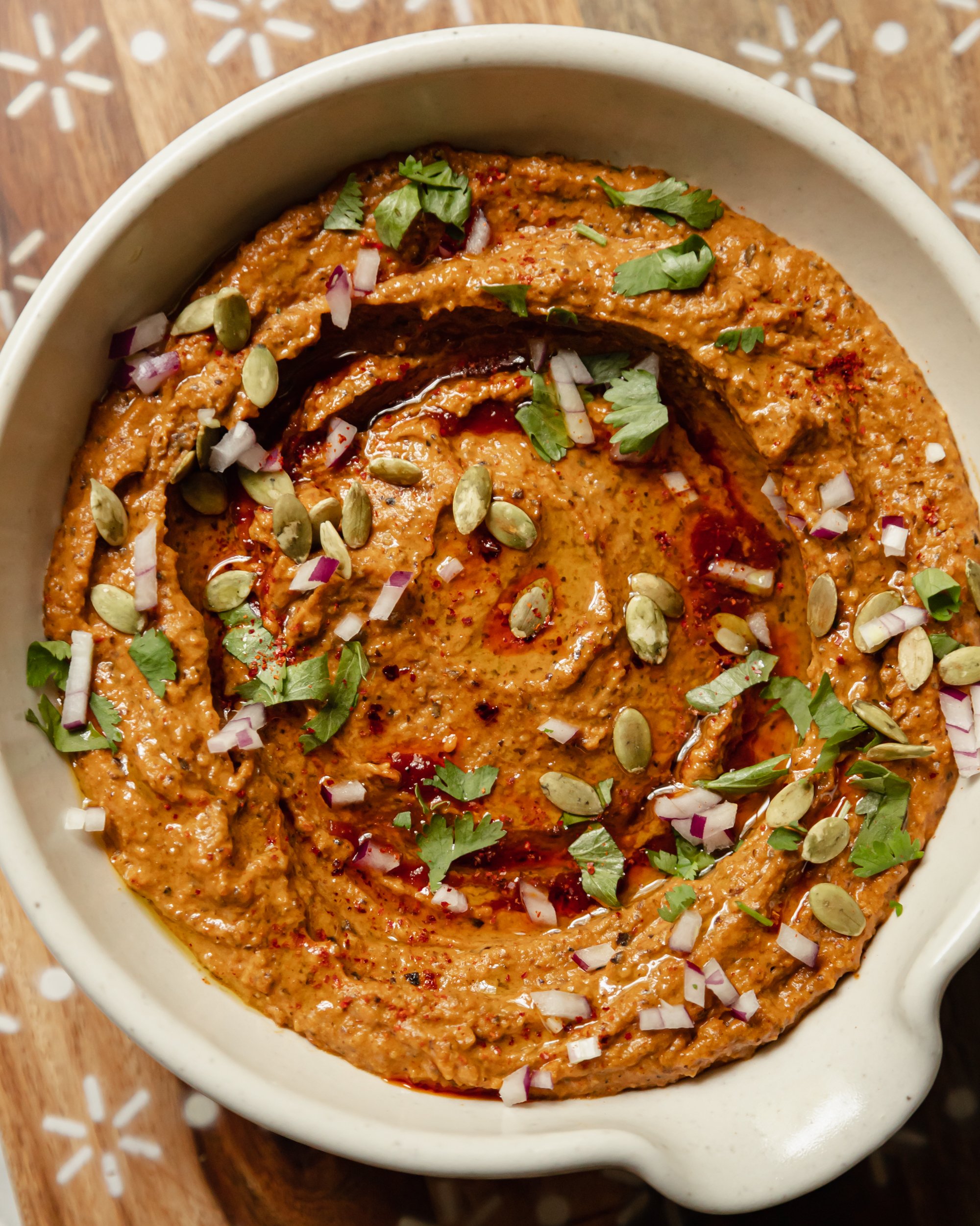 An up close, overhead shot of a deep red black bean dip that is garnished with pumpkin seeds, finely diced red onion, olive oil, and cilantro.