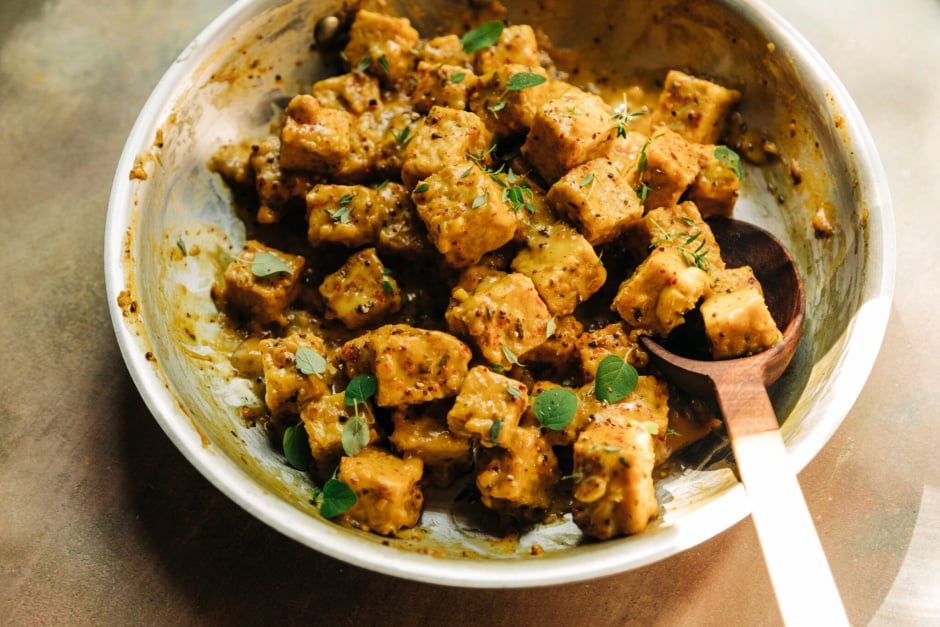 A 3/4 angle shot of saucy chunks of tempeh in a skillet. The sauce is golden yellow and the tempeh is garnished with fresh thyme and oregano leaves.