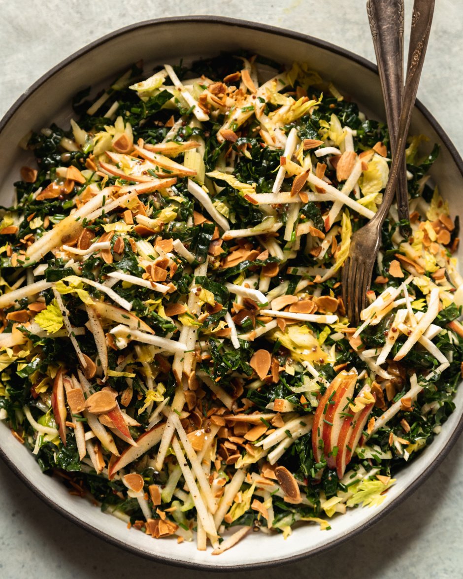An up close, overhead shot of an apple kale salad with toasted almonds, photographed in a white ceramic dish that is rimmed with matte black.