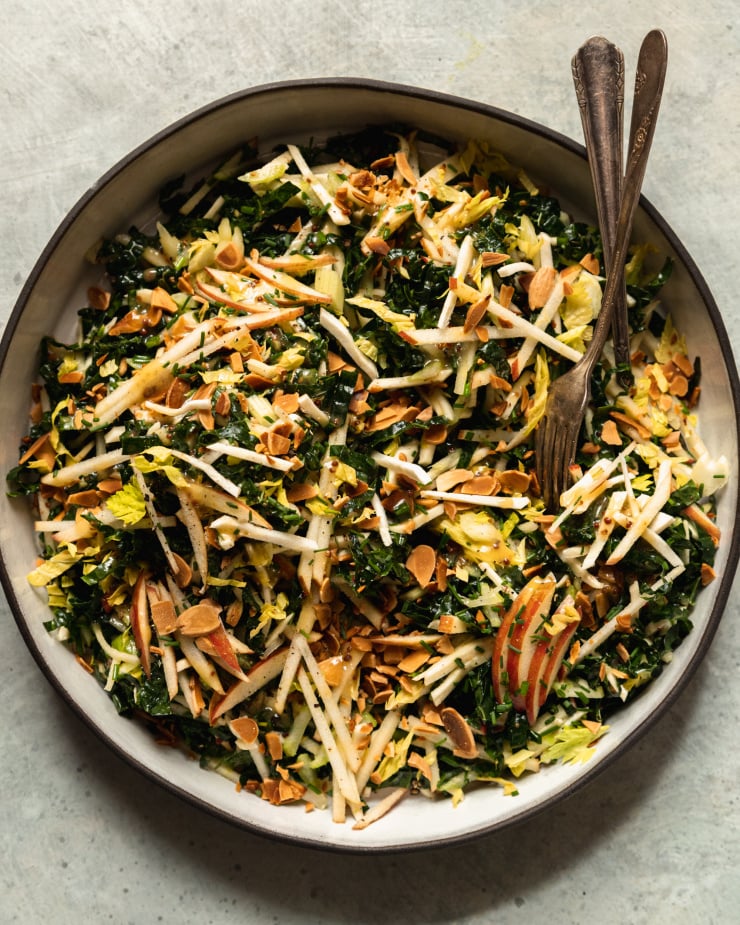 An overhead shot of an apple kale salad with toasted almonds, photographed in a white ceramic dish that is rimmed with matte black.