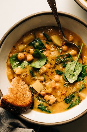 An overhead shot of a creamy and thick lemony chickpea soup with spinach and potatoes in a white ceramic bowl edges with grey. A piece of whole grain bread is sticking out of the soup.