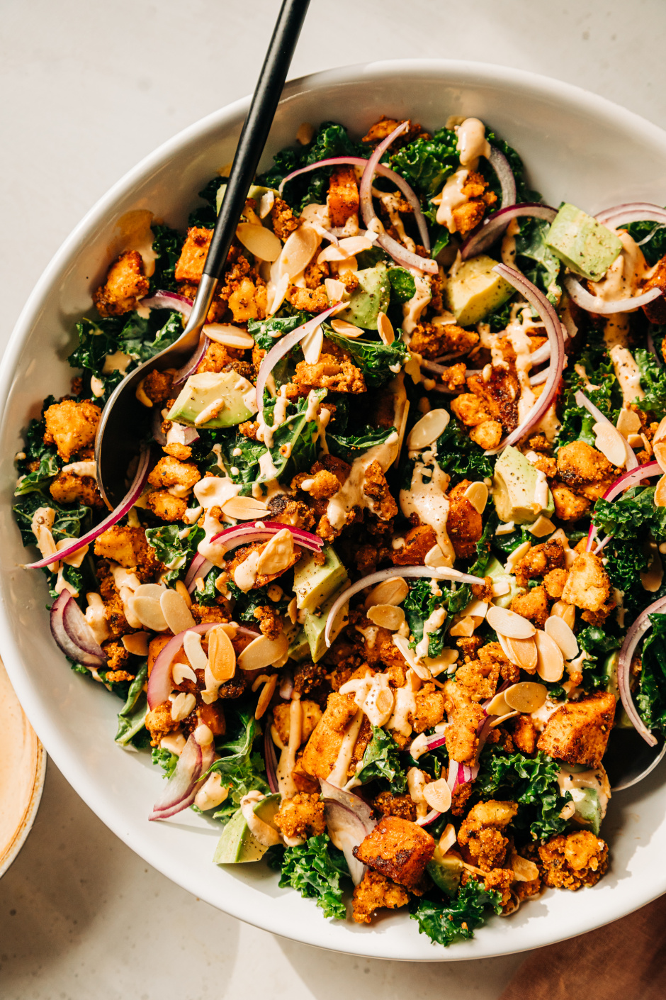 An overhead shot of a kale salad with creamy dressing on top, crispy bits of tofu, chunks of roasted sweet potato, slices of red onion, and cubes of avocado. The salad is in a white bowl.