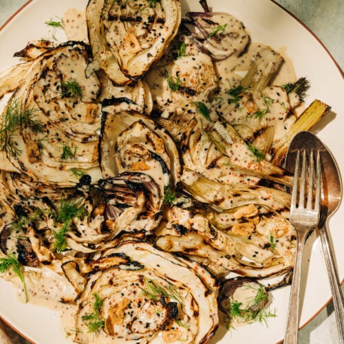 An overhead shot of grilled cabbage and fennel in direct sunlight. The vegetables are covered in a creamy mustard seed sauce and garnished with fennel fronds.