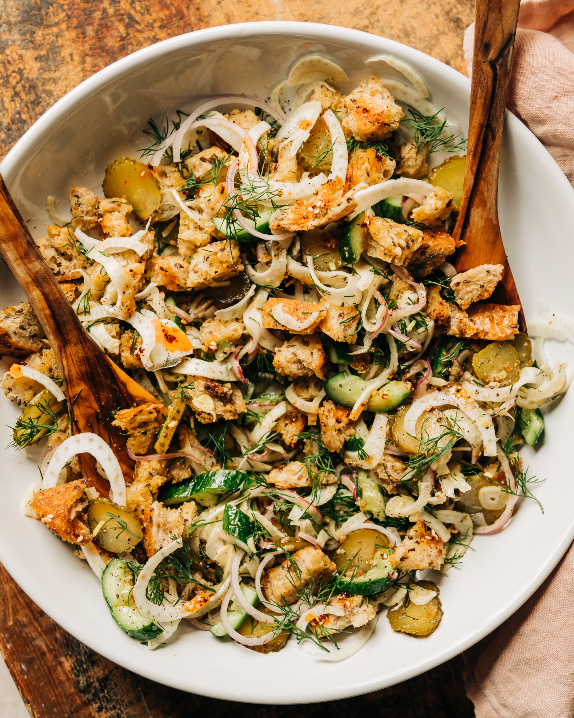 An overhead shot of a pickle panzanella salad with crispy bits of bread, chopped cucumbers, pickles and red onion, plus shaved fennel and fresh dill. Photographed in a wide, white bowl with wood serving tongs. All set on a worn wood background.