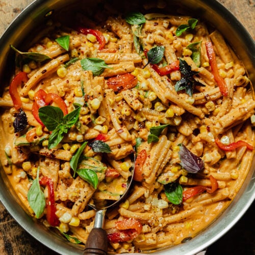 An overhead shot shows a creamy vegan red pepper pasta in a stainless steel pan. The short-shaped pasta is garnished with fresh basil leaves, strips of roasted red pepper, charred corn kernels, dried chili flakes, and freshly ground pepper.