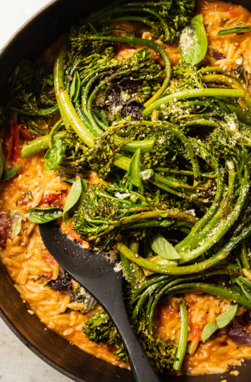 An up close overhead shot of a creamy vegan tomato orzo dish in a pot with roasted broccolini and fresh basil on top.