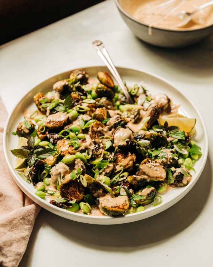 A slight 3/4 angle shot of a plate of crispy roasted brussels sprouts and cooked edamame beans, drizzled with a creamy peanut sauce and finished with chopped green onions, mint, and sesame seeds.