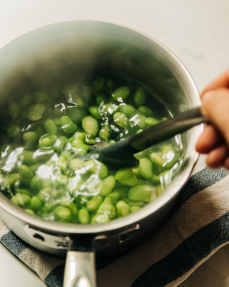 A hand is seen stirring a steaming pot of cooked edamame beans against a light grey background.