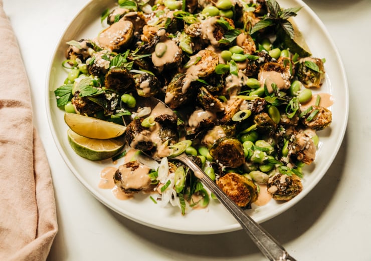 An up close, overhead shot of a plate of crispy roasted brussels sprouts and cooked edamame beans, drizzled with a creamy peanut sauce and finished with chopped green onions, mint, and sesame seeds.