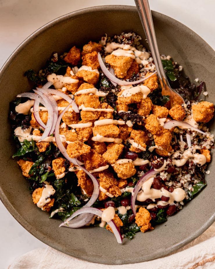 An overhead shot of a grain bowl with quinoa, kale, black beans, thinly sliced red onion, and crispy baked tofu bits. A creamy sauce is drizzled on top and a fork is sticking out of the bowl.