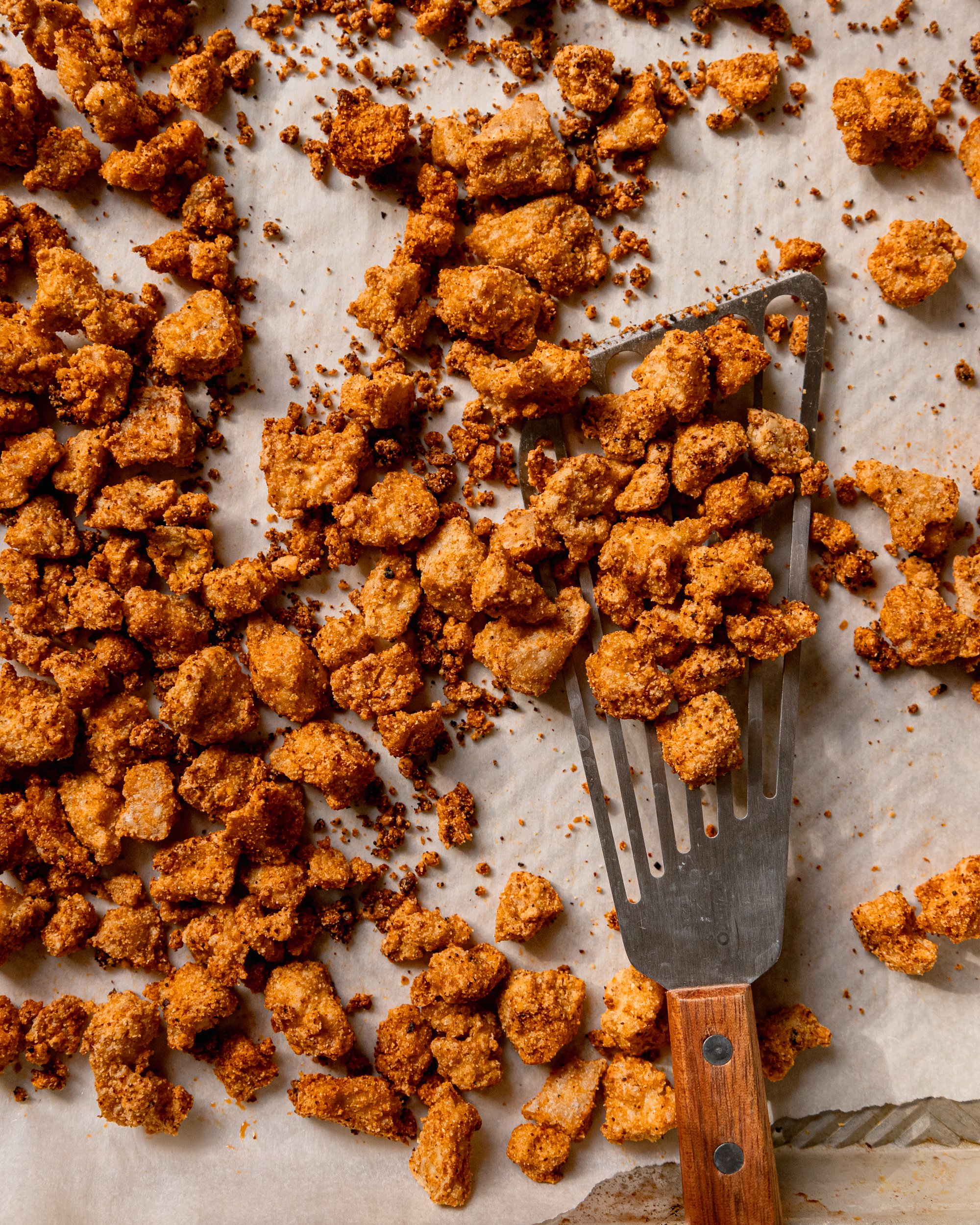 An overhead shot of a baking sheet filled with crispy baked tofu bits. A wooden handled fish spatula is lifting some of the pieces off of the baking sheet.