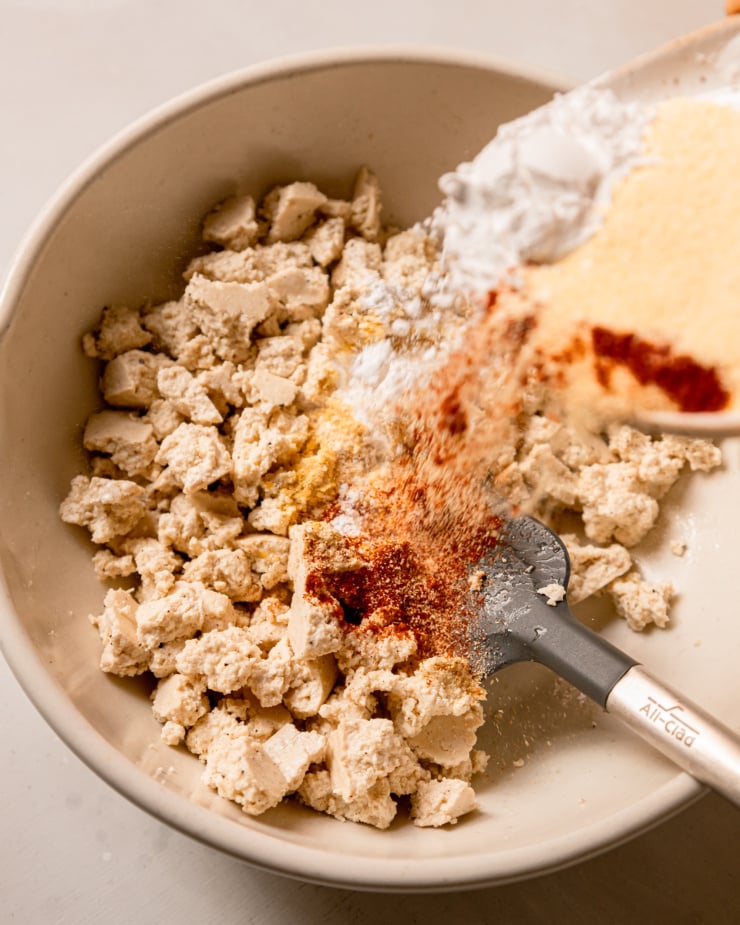 An overhead shot shows a bunch of spices, arrowroot, and cornmeal being poured on top of a bowl containing torn pieces of tofu.