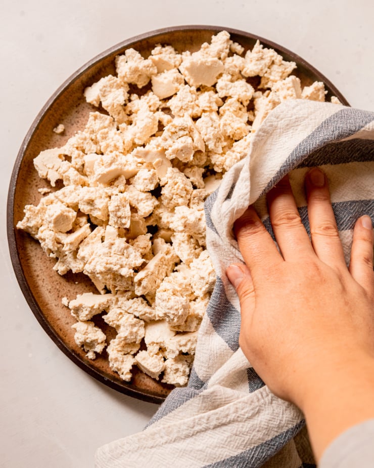 An overhead shot shows a hand using a kitchen towel to blot pieces of tofu dry on a plate.