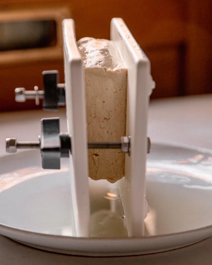 A head on shot shows a 1 lb piece of tofu being pressed in a tofu press set over a bowl to catch the water.