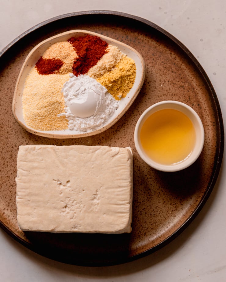 An overhead shot shows a 1 lb piece of tofu, a small plate with spices, arrowroot starch and cornmeal, and a small bowl of avocado oil; all on top of a round, brown plate.