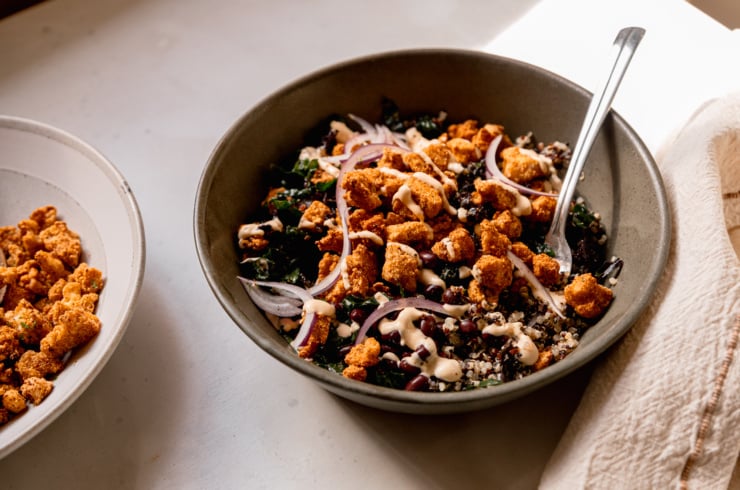 A 3/4 angle shot of a grain bowl with quinoa, kale, black beans, thinly sliced red onion, and crispy baked tofu bits. A creamy sauce is drizzled on top and a fork is sticking out of the bowl.