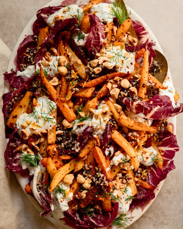 An up close, overhead shot of a harissa carrot and parsnip salad with cooked quinoa, chopped fresh dill, a creamy vegan yogurt sauce dolloped on top, and chopped toasted hazelnuts. All of this is set on a bed of radicchio leaves in a platter