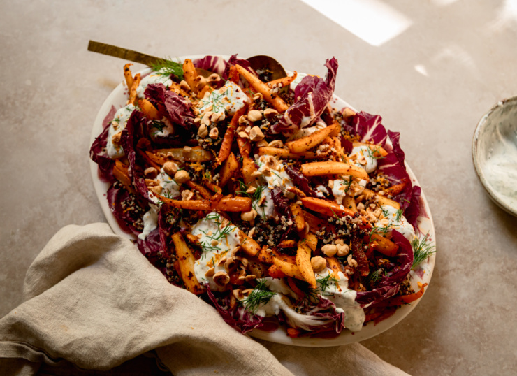 An 3/4 angle shot of a harissa carrot and parsnip salad with cooked quinoa, chopped fresh dill, a creamy vegan yogurt sauce dolloped on top, and chopped toasted hazelnuts. All of this is set on a bed of radicchio leaves in a platter