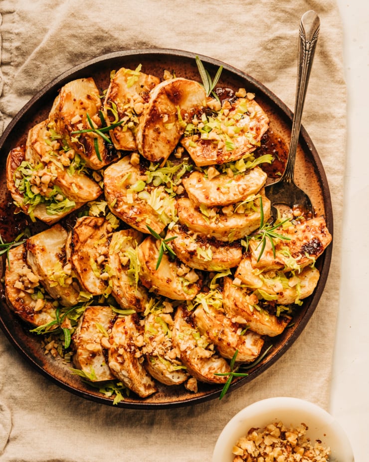 An overhead shot of cider glazed celery root pieces fanned out on a speckled brown plate. The celery root is topped with chopped walnuts, rosemary leaves, and finely chopped celery heart leaves.